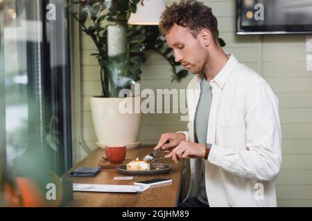 Seitenansicht eines lockigen Mannes, der Besteck in der Nähe von Zitronentherb auf Teller, Smartphone und Tasse im Café hält Stockfoto