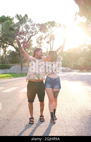Junge, coole tätowierte Paare brüllen, erheben ihre Arme und lachen mit einem Skateboard. Stockfoto