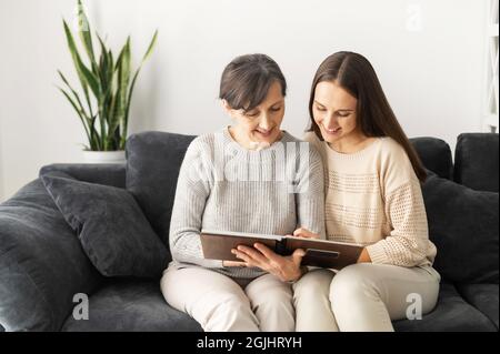 Zwei Frauen unterschiedlicher Generationen verbringen Freizeit zu Hause, eine ältere Mutter und eine Erwachsene Tochter schauen sich gemeinsam Bilder im Fotoalbum an, erinnern sich an angenehme Momente. Familienbande Stockfoto