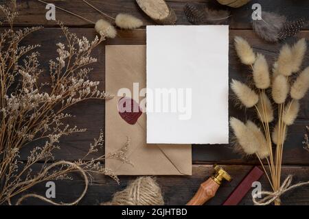 Leere Papierkarte auf versiegeltem Umschlag und Holztisch mit getrockneten Pflanzen, Draufsicht. Boho-Mock-up-Szene mit Grußkartenvorlage Stockfoto