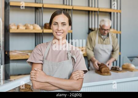 Porträt einer glücklichen jungen Brünette in Schürze, die mit gekreuzten Armen in einer Bäckerei ihres Vaters steht Stockfoto