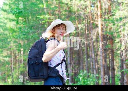 Junge Touristenfrau mit einem Rucksack in einem Strohhut im Wald vor dem Hintergrund von grünen Bäumen an einem hellen Sommersonnentag. Selektiver Fokus Stockfoto