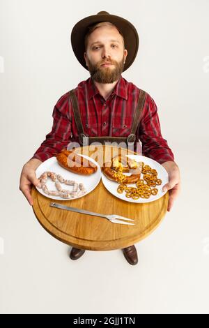 Draufsicht auf den jungen bärtigen Mann, Kellner in traditioneller bayerischer Tracht mit rundem Holztablett mit festlichen Speisen, Snacks, Würstchen und Brezeln Stockfoto