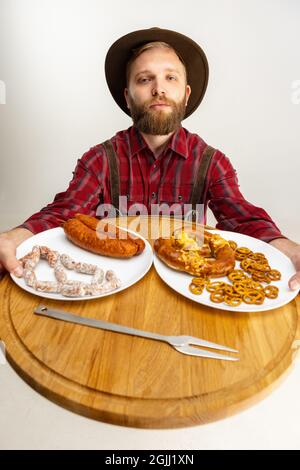 Draufsicht auf den jungen bärtigen Mann, Kellner in traditioneller bayerischer Tracht mit rundem Holztablett mit festlichen Speisen, Snacks, Würstchen und Brezeln Stockfoto