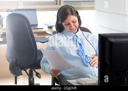Eine Schwangerin arbeitet im Büro am Computer. Der Mitarbeiter sitzt auf einem Stuhl gegenüber dem Fenster und dreht sich um. Stockfoto