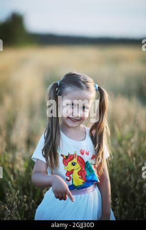 Ein Mädchen mit zwei Schwänzen, mit einem Einhorn im Fußball gekleidet, geht auf einem Weizenfeld spazieren und lächelt. Stockfoto