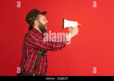 Komisches Porträt eines bärtigen Mannes, eines Kellners im Hut und einer traditionellen bayerischen Tracht, das auf einem roten Studiohintergrund isoliert auf ein Megaphon schreit. Oktoberfest Stockfoto