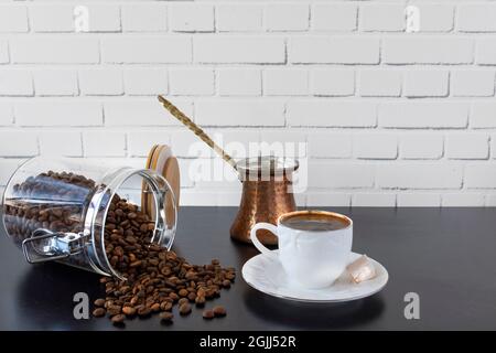 Vor einer weißen Backsteinmauer, auf schwarzem Hintergrund, eine Tasse türkischer Kaffee, türkischer Genuss, Kanne und ein Glas Bohnen. Selektiver Fokus. Stockfoto