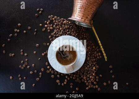 Türkischer Kaffee auf einer Tasse Rauch auf schwarzem Hintergrund. Verstreute Bohnen und ein umgedrehter Topf. Stockfoto