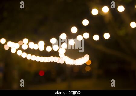 Verschwommener abstrakter Hintergrund, Hinterhofbeleuchtung, Licht im Abendgarten, elektrische entkochte Girlande. Lampe Girlande von Glühbirnen auf Bäumen beleuchten Nacht Szene Stockfoto