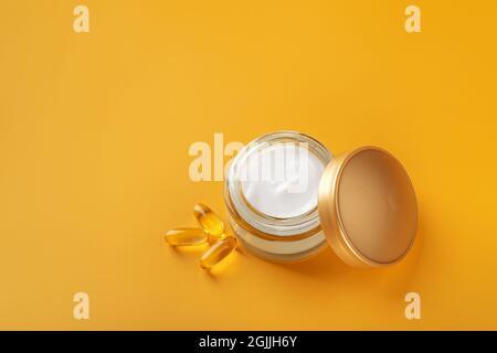 Feuchtigkeitsspendende Hautpflegecreme in einem offenen Glas mit goldenem Deckel und wenigen ätherischen Ölkapseln vor hellgelbem Hintergrund. Gesichts- und Körperpflege. Stockfoto