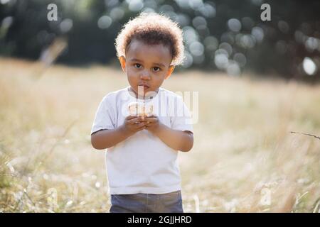 Porträt eines hübschen afroamerikanischen Jungen in weißem T-Shirt und blauer Jeans mit süßem Cupcake in den Händen, während er über die Natur im Sommer posiert. Sonnenlicht in lockig dunklem Haar. Stockfoto
