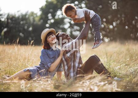 Schöne kaukasische Frau und schöner afroamerikanischer Mann, der während des Picknicks im Freien mit ihrem kleinen Sohn spielt. Fröhliche dreiköpfige Familie, die auf Gras sitzt und Spaß zusammen hat. Stockfoto