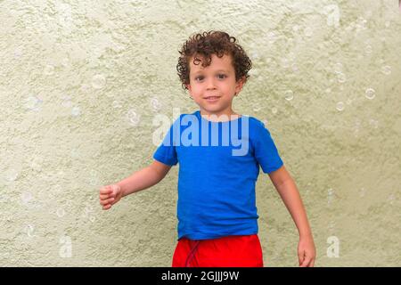 Netter und hübscher Junge mit lockigen Haaren und kaukasischem Aussehen, der die Kamera, umgeben von Seifenblasen, anschaut Stockfoto