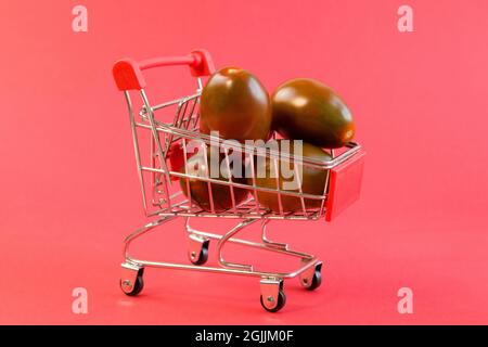 Nahaufnahme eines kleinen Einkaufswagens mit frischen schwarzen Tomaten auf hellrotem Hintergrund Vorderansicht Stockfoto