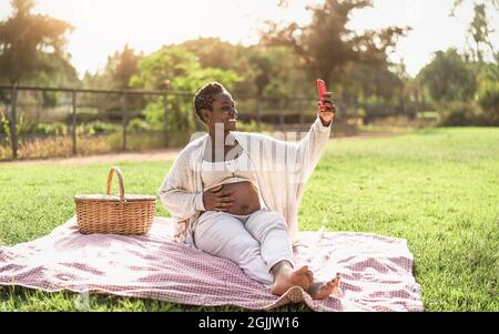 Afrikanische Schwangere, die Selfie mit dem Handy macht, während sie ein Picknick im Park macht - Mutterschaft Lifestyle-Konzept Stockfoto