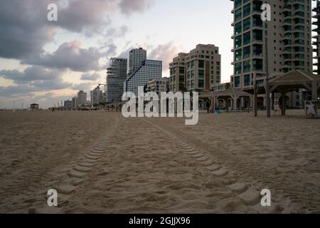 Reifenspuren, die im Morgengrauen am Strand von Tel Aviv, Israel, im Sand hinterlassen wurden. Hotels im Hintergrund. Stockfoto