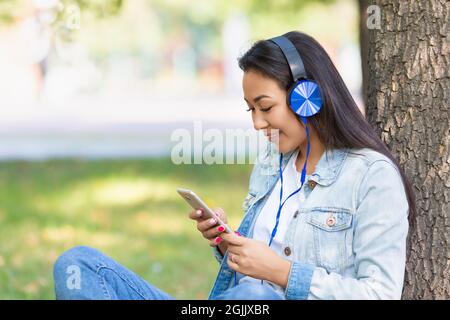 Foto einer asiatischen Frau in Kopfhörern mit Smartphone. Sie lächelt und hört sich ihre Lieblingsmusik oder ihre Unterrichtsstunde an. Stockfoto