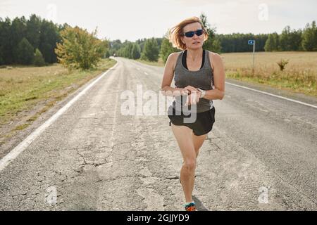 Reife sportliche Frau in Sportkleidung und in Sonnenbrillen, die entlang der Landstraße laufen Stockfoto