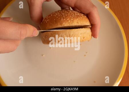 Frau schneidet Hamburgerbrötchen mit Sesamsamen und halbfertig geschnittenes Messer Stockfoto