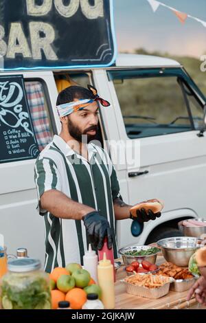 Junger bärtiger Mann, der am Strand Fastfood mit einem Café-Van im Hintergrund zubereitete Stockfoto