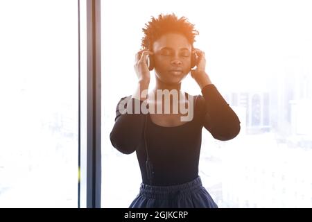 Eine afroamerikanische Frau hört Musik mit Kopfhörern. Eine junge Frau in einer Sportuniform auf dem Hintergrund eines Fensters. Hochwertige Fotos Stockfoto