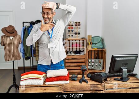 hispanischer Mann mittleren Alters, der als Manager in der Einzelhandelsboutique arbeitet, lächelt fröhlich und guckst einen Buh mit Händen, die Gesicht zeigen. Überrascht und begeistert Stockfoto