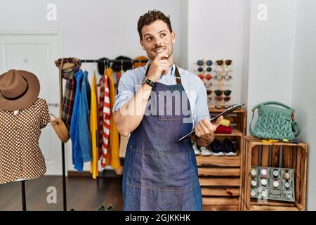 Hübscher junger Mann, der als Manager in der Einzelhandelsboutique arbeitet, mit der Hand am Kinn, der über Fragen nachdenkt, nachdenklichen Ausdruck. Lächelnd und nachdenkliches Gesicht. d Stockfoto
