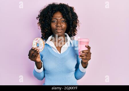 Junge afroamerikanische Frau essen Donut und trinken Kaffee ahnungslos und verwirrt Ausdruck. Zweifel Konzept. Stockfoto