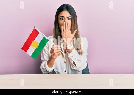 Junge hispanische Frau mit der Flagge kurdistans auf dem Tisch sitzend, die den Mund mit der Hand bedeckt, schockiert und Angst vor Fehlern. Überraschter Ausdruck Stockfoto