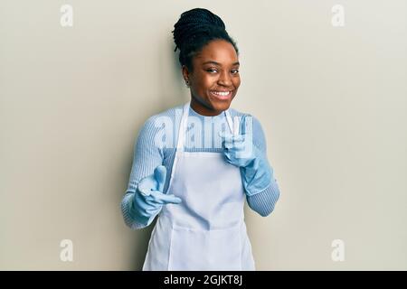 afroamerikanische Frau mit geflochtenen Haaren trägt Reinigungsschürze und Handschuhe zeigen Finger zur Kamera mit glücklichen und lustigen Gesicht. Gute Energie und Vibes. Stockfoto