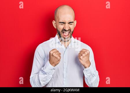 Junger kahlköpfiger Mann in eleganten Kleidern, der überrascht und erstaunt über den Erfolg mit erhobenen Armen und geschlossenen Augen zelebriert Stockfoto