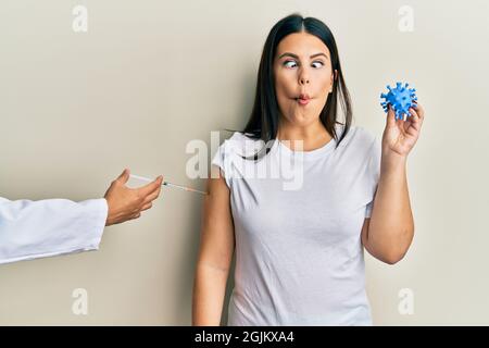 Schöne Brünette Frau bekommen Impfstoff hält Coronavirus Spielzeug machen Fisch Gesicht mit Mund und blinzeln Augen, verrückt und komisch. Stockfoto