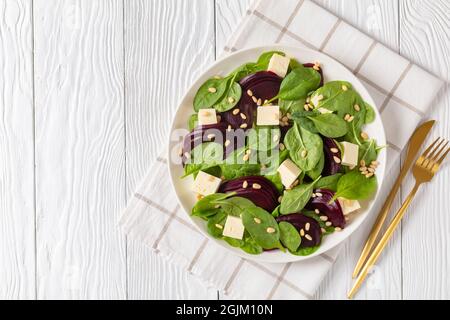 Gerösteter Rübensalat mit gewürfeltem Feta-Käse, Pinienkernen und Spinat auf einem weißen Teller mit goldenem Besteck auf einem Holztisch, kanadische Küche, flaches Lay, f Stockfoto