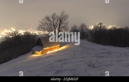 Fantastische Winterlandschaft mit glühendem Holzhaus vor dem Hintergrund glühender Lichter der Stadt im Nebel. Gemütliche Hütte in den Karpaten. Weihnachtsfeiertagskonzept Stockfoto