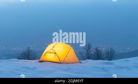 Gelbes Zelt von innen beleuchtet vor dem Hintergrund von leuchtenden Stadtlichtern im Nebel. Erstaunliche verschneite Landschaft. Touristen campen in den Winterbergen. Reisekonzept Stockfoto