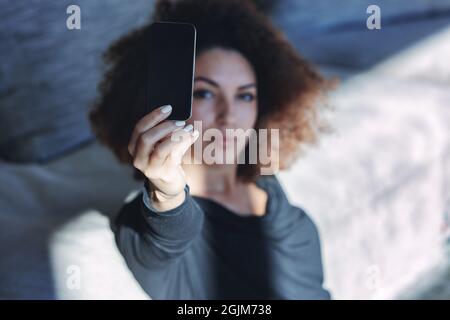 Selbstbewusste junge Frau, die ein Selfie auf ihrem Mobiltelefon macht, während sie sich auf dem Boden entspannt und sich auf ein Ofa stützt, mit Fokus auf das Smartphone Stockfoto