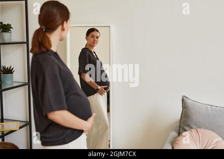 Schöne Schwangeren junge Frau in bequemer Kleidung stehen in gemütlichen Wohnzimmer und Blick auf Bauch im Spiegel Stockfoto