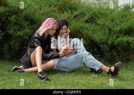 Zwei Teenager-Mädchen beobachten etwas auf einem Smartphone, auf dem Gras hinter den Büschen im Park Stockfoto