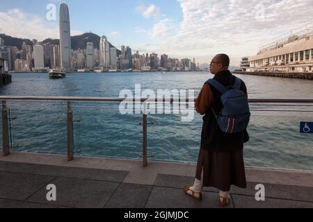 Ein Mönch wartet auf die Star Ferry am Pier in Tsim Sha Tsui mit dem Wolkenkratzer des International Finance Center dahinter Stockfoto