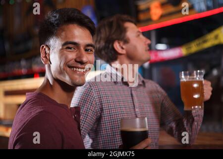 Ein hübscher junger Mann lächelt zur Kamera, während er mit einem Freund Bier trinkt Stockfoto