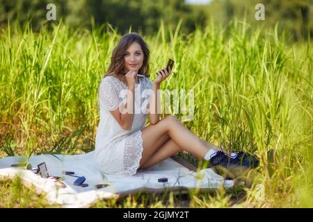Junge blonde Frau in weißem Kleid sitzt auf einem Picknicktuch im hohen Gras. Weibliche Make-up-Künstlerin mit Make-up-Accessoires verteilt um sie herum. Stockfoto