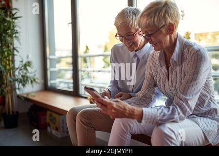 Ältere weibliche Kollegen schauen sich Smartphones an, während sie in entspannter Atmosphäre am Arbeitsplatz eine Pause einlegen. Unternehmen, Büro, Job Stockfoto