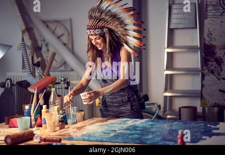 Eine junge Künstlerin mit einer Kriegshaube, die in angenehmer Atmosphäre im Atelier an einem neuen Gemälde arbeitet. Kunst, Malerei, Atelier Stockfoto