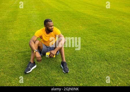 Junger erwachsener Mann, der nach dem Training in stilvoller Sportkleidung auf dem Rasen sitzt, hält ein Smartphone und entspannt. Sportlicher afroamerikanischer Rüde, der eine Pause einnahm Stockfoto