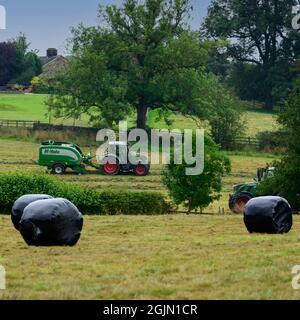 Heu- und Silageherstellung (Landwirt im landwirtschaftlichen Traktor bei der Arbeit in ländlichen Feldpressen, sammeln Wrapping trockenes Gras in runden Ballen) - Yorkshire England Großbritannien Stockfoto