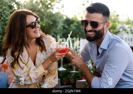 Fröhliche junge Leute, die bei einer Gartenterrasse Spaß mit lachenden, klirrenden tropischen Cocktails haben. Freunde toasten im Freien Stockfoto