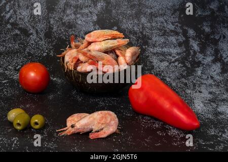 Frische Paprika, Tomaten, Oliven und gefrorene Garnelen in einer umweltfreundlichen Kokosnussschale auf dunklem Grund Stockfoto