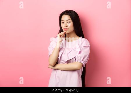 Hmm, interessant. Nachdenklich und gerissen asiatische Mädchen mit Idee, planen, etwas zu tun, Blick auf Kamera und berühren Kinn mit nachdenklichen Gesicht, stehend Stockfoto