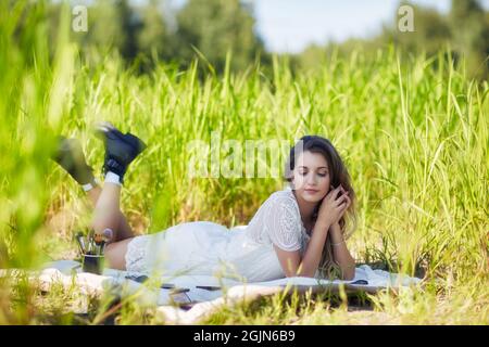 Junge blonde Frau in weißem Kleid liegt auf einem Picknicktuch im hohen Gras. Weibliche Make-up-Künstlerin mit Make-up-Accessoires verteilt um sie herum. Stockfoto